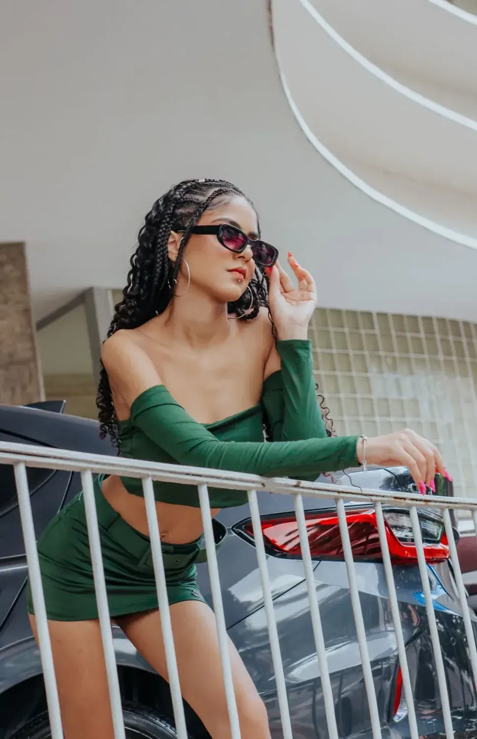 Fashionable woman in green outfit leaning on railing, showcasing urban style with sunglasses.