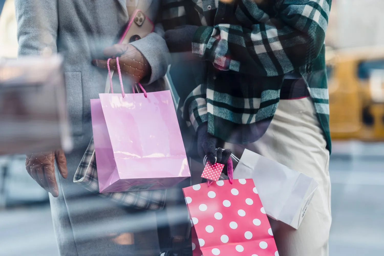 Crop faceless women in trendy outerwear carrying paper bags in hands and standing near shop window