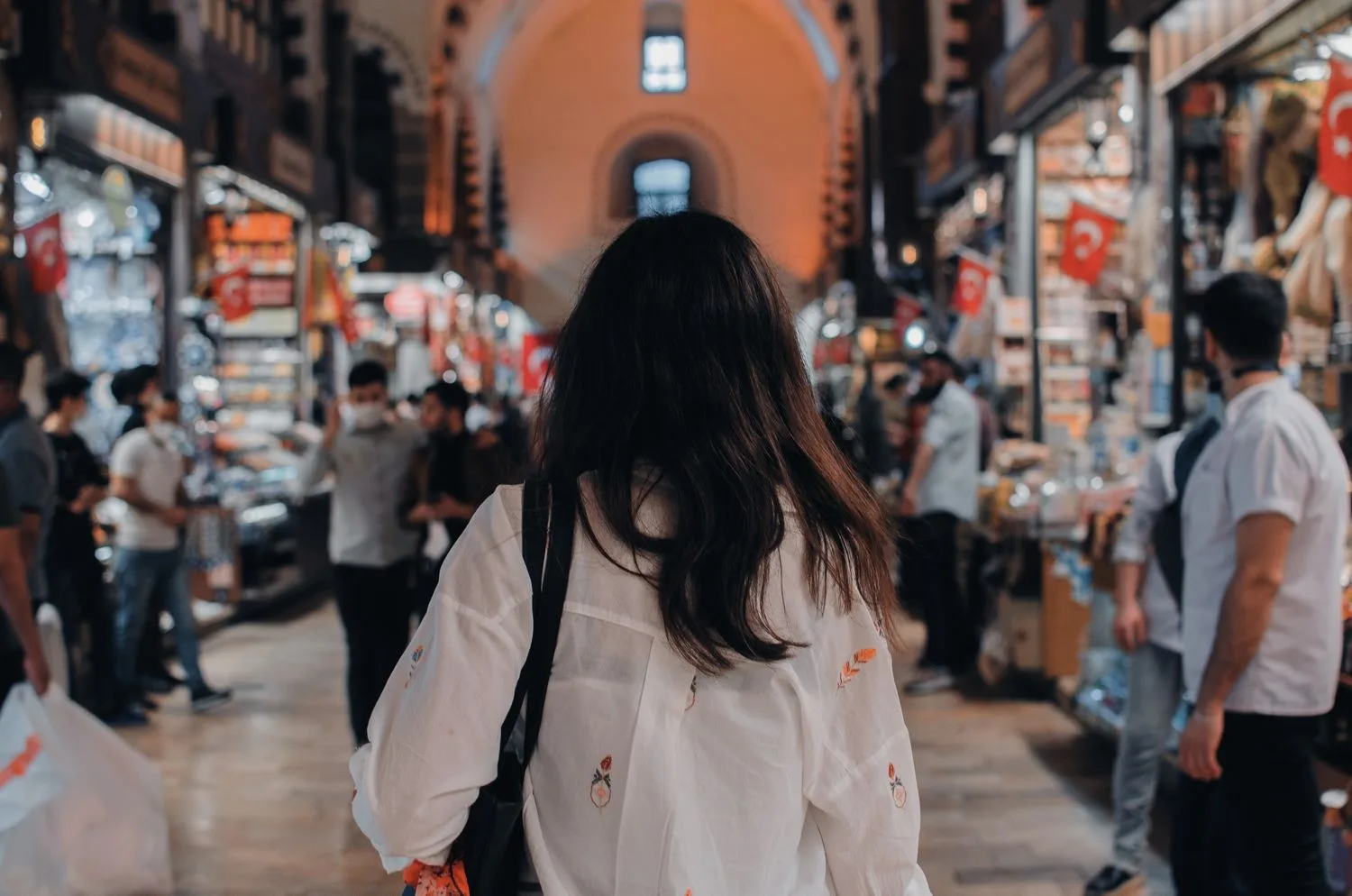 Back view of unrecognizable female with long black hair wearing white shirt walking through alley of shops in Turkey